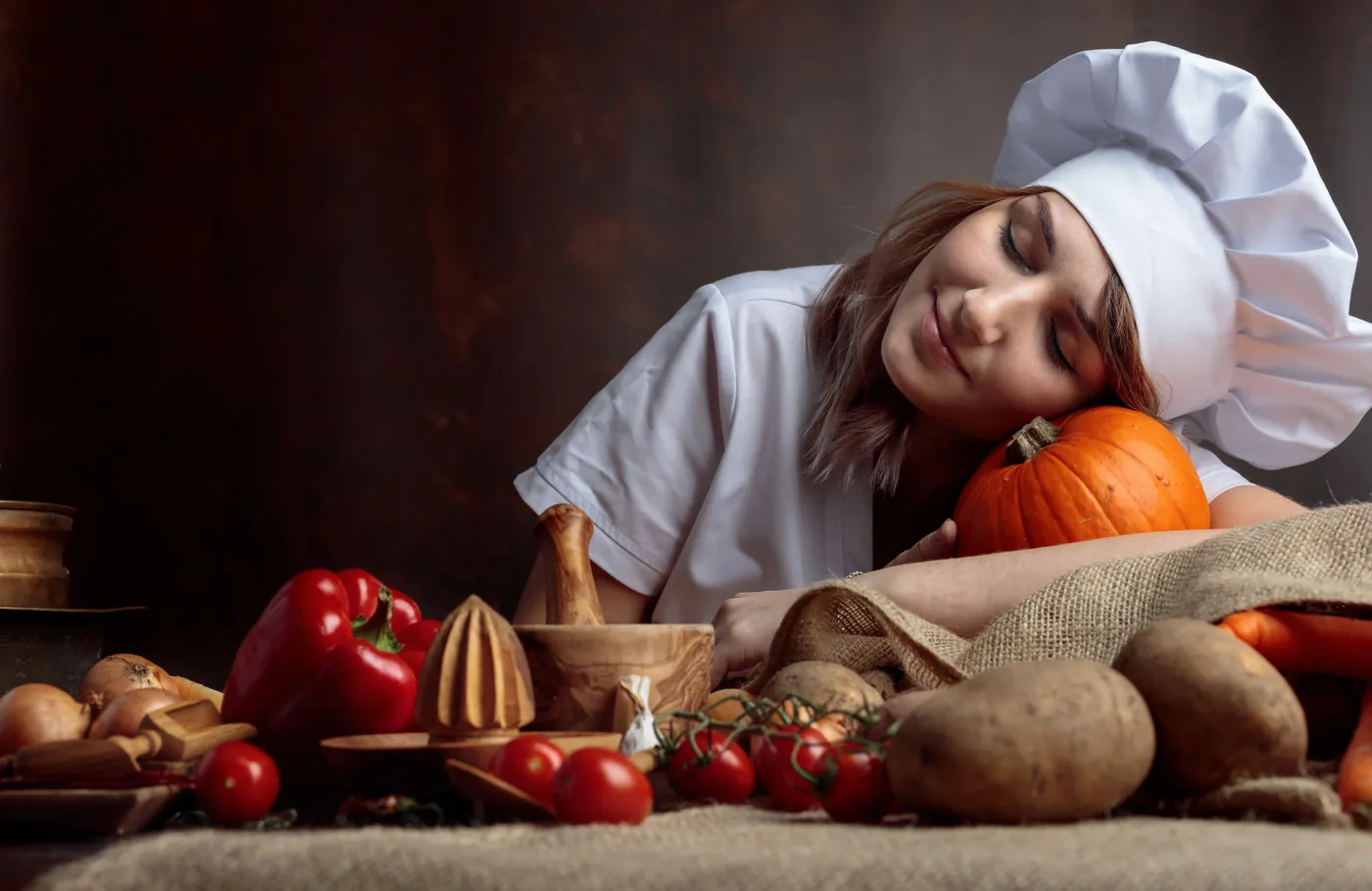 A smiling chef resting her head on a pumpkin beside fresh vegetables